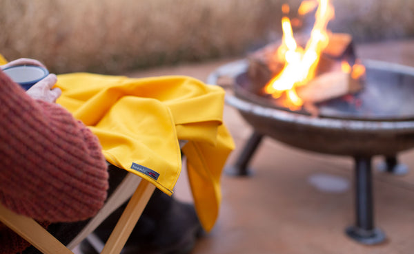 A person sitting in front of a fire pit, holding a cup of hot tea with a Saffron Shepherd's Wool draped over their lap for warmth.